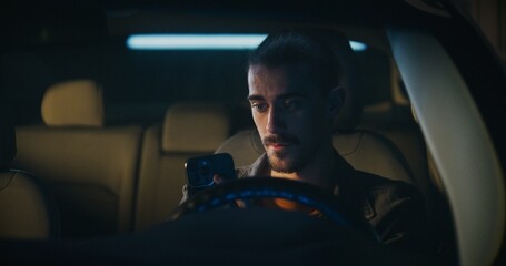 Obraz premium Seated in Driver Seat, Serious Male With Man Bun and Mustache Firmly Holds Smartphone, Typing Detailed Message. Man Concentrated, Resolve and Purpose. Private Moment of Modern Mobile Interaction.