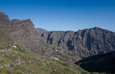 Panoramic view of Masca village in Teno Rural Park, Tenerife, Spain. Dramatic volcanic cliffs surround the small settlement nestled deep in a rugged mountain gorge under clear blue skies
