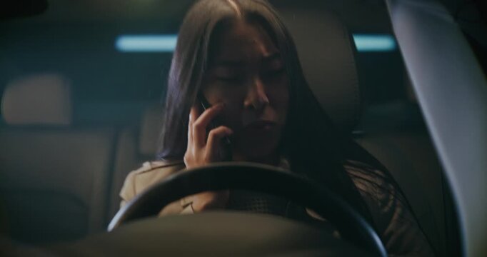 Settles in Driver Seat, Asian Woman Already Engaged in Tense Smartphone Conversation, Face Clouded With Worry. Female Leans Against Headrest, Sharing Details of Difficult Day With Person on Other End.