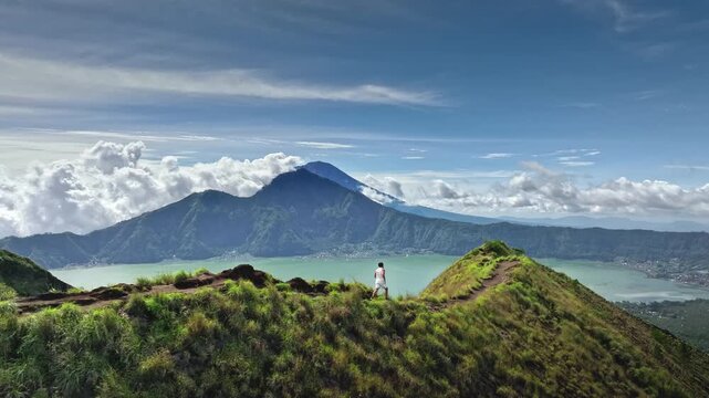 Aerial view sportsman running along narrow green ridge of Mount Batur with panoramic view of Lake Batur and lush volcanic peaks under a clear blue sky. Active sport and recreation. Slow motion shot