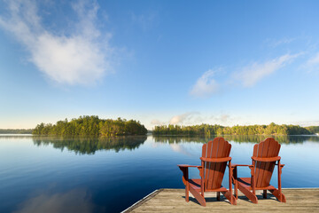 Fototapeta premium Two Adirondack chairs on a wooden dock overlooking a calm, misty lake with forested islands and a blue morning sky. Ample copy space in sky and water.