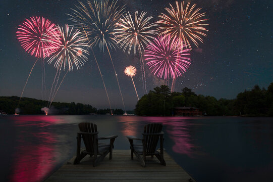 Colorful fireworks exploding in the night sky over a calm lake, with two chairs silhouetted on a dock in the foreground. Ample copy space.