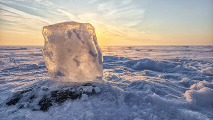 A translucent ice block backlit by a golden sunset on a vast, snowy plain. This serene winter landscape captures the cold, peaceful mood of the arctic with warm light and a pastel sky.