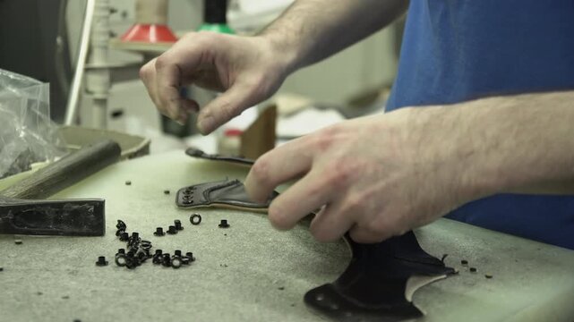 Skilled artisan meticulously attaches small metal grommets by hand to dark leather material for custom footwear production in a busy workshop setting