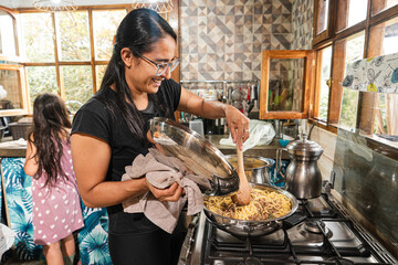 Mother mixing pasta with sauce in cozy kitchen while daughter stands nearby. Authentic family cooking moment. © JulianSarmiento