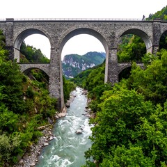 Fototapeta premium Stone arched bridge spans a river, surrounded by lush green trees and a distant mountain peak under cloudy sky