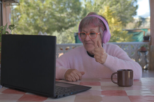 Older woman with headphones happily waving while looking at her laptop in her country house