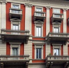 Obraz premium Colorful historic building facade in Milan, Italy with red walls, classical columns and decorative balconies showcasing elegant European architecture and symmetry.