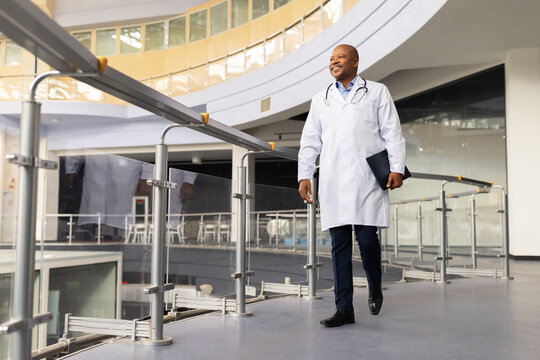 Mature black doctor walking in modern hospital atrium with folder, confident african american healthcare professional in bright contemporary interior, copy space