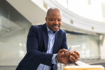 Fototapeta premium Thoughtful mature black businessman looking at smartphone in office atrium, confident corporate leader reviewing information in modern architectural setting