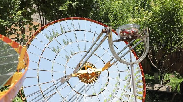 A large parabolic solar cooker is set outdoors in a garden. The mirrored dish concentrates solar energy to cook food inside a glass container at its focal point.