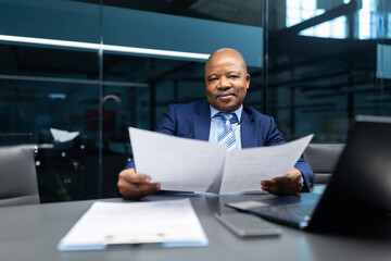 Fototapeta premium Confident mature black businessman reviewing documents while sitting at desk with laptop in modern office. Executive analyzing paperwork and business reports