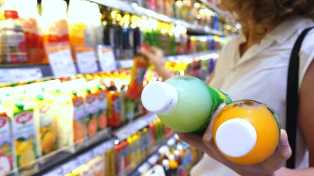 Shopper holding orange and lime juice bottles while choosing another healthy beverage from a refrigerated shelf in a grocery store, demonstrating a focus on conscious consumerism and nutrition