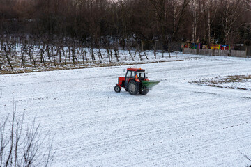 Red tractor with spreader working on snow covered agricultural field in winter, farming machinery fertilizing soil during cold season, rural landscape with industrial equipment.