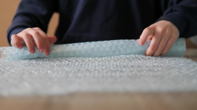Close-up of hands rolling and folding bubble wrap in 4K. Detailed footage of a boy manipulating plastic packing material on a wooden surface, ideal for themes of shipping, e-commerce, and sensory play