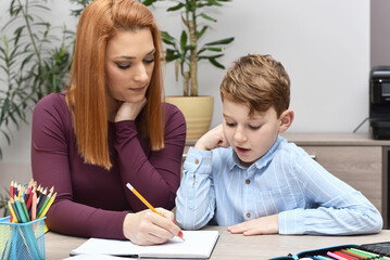Mother helping her young son with homework at home. Woman and child sitting at a desk, writing in a notebook together. Concept of education, parenting, learning, and family support.