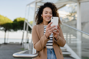 Fototapeta premium pretty curly smiling woman walking in city street in stylish jacket, using smartphone