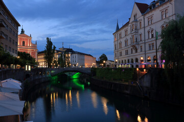 Fototapeta premium Blue hour view in Ljubljana, where a magical twilight light softens historic buildings and reflects on the river, blending deep blues with warm city glows at dusk.