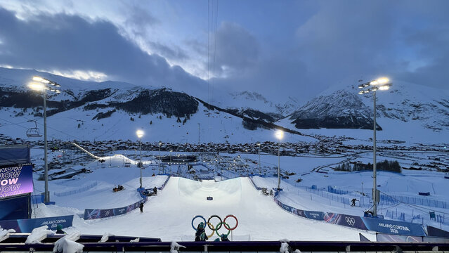 Livigno, Italy - February 20, 2026: Milano Cortina 2026 Olympic rings installation with snowy halfpipe venue and illuminated village panorama in Alps.