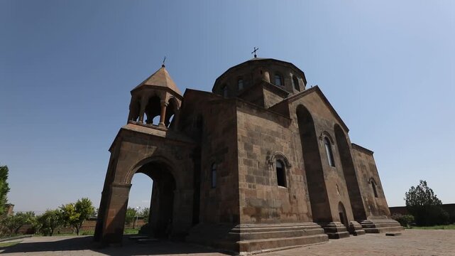 Wide shot of a historic, ancient stone church featuring a central dome and an attached bell tower, standing in a paved courtyard against a clear blue sky.

