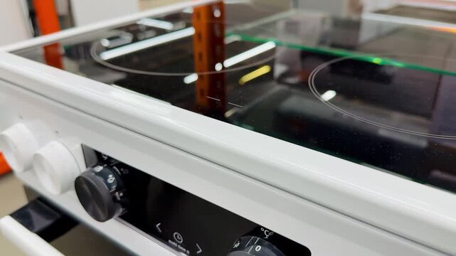 White electric stove with glass cooktop on showroom shelf, visible control knobs and mode dials, crisp enamel body, reflective.