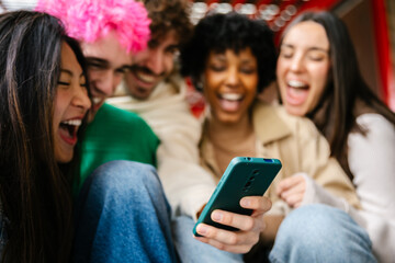 Happy multiracial friends are sitting outdoors on university steps, laughing while watching mobile...