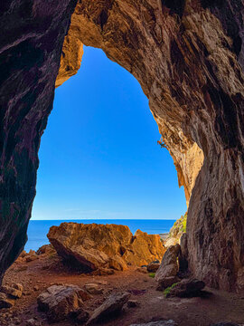 LOW ANGLE VIEW: Strong female climber tackles steep limestone tufas at the entrance of Grotta del Cavallo. Deep blue Mediterranean Sea and sport climbing action is framed by a dramatic cave arch.