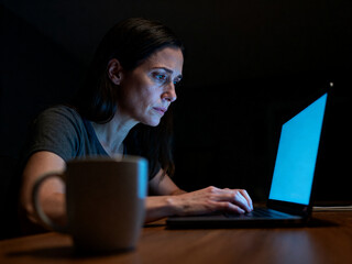 Woman working on laptop in dark room at night with focused expression and coffee cup on table