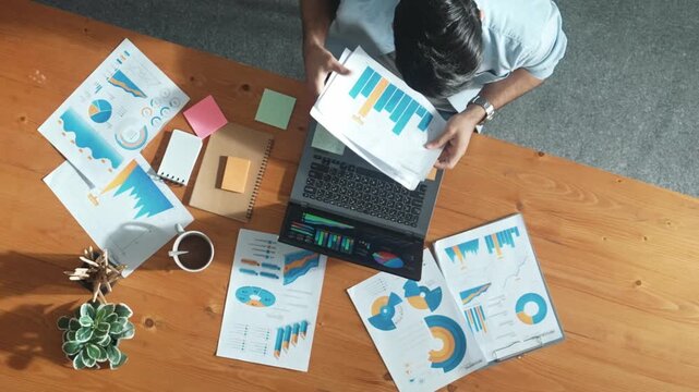 Top aerial view of manager working on laptop while looking at data analysis. Leader compare financial statistic graph while making decision at meeting table with document scatter around. Convocation.
