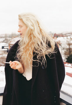 Woman with long blonde hair holding a cigarette on a snowy rooftop 