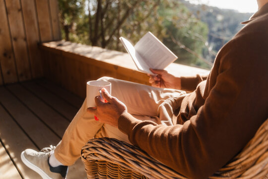 Man Relaxing with Tea and Book on Log Cabin Balcony during Sunny Day
