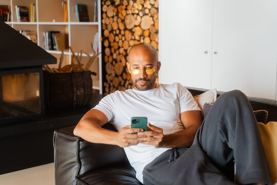 Man Relaxing with Collagen Patches, Texting on His Sofa at Home