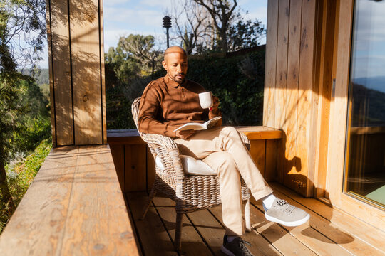 Bald Man Reading Book and Drinking Tea on Log Cabin Balcony