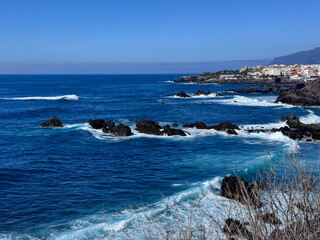 Blue ocean with waves crashing against the rocks along the coastline of Los Gigantes, near cliffs with blue sky