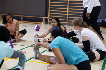 Diverse female teenage volleyball players stretching in circle on gym floor with volleyballs
