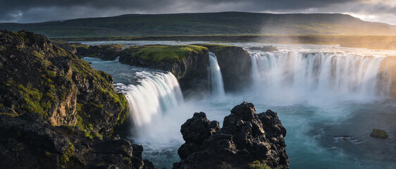 Panoramic view of Godafoss waterfall in Iceland with powerful cascading water, misty spray, dark volcanic rock formations, lush green moss, and dramatic golden sunset light over the horizon.