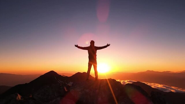 Silhouette of a triumphant man standing with arms outstretched on a rocky mountain peak at sunset with warm, vibrant colors.