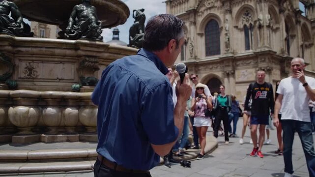 Tour guide speaking near cathedral fountain, caucasian man holding microphone addresses busy historic square filled with walking tourists, backpacks, cameras, sunlit stone architecture and ornate