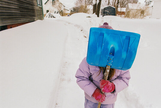Child in winter clothes covers her face with a snow shovel 