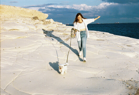 Woman walks small dog along a sandy beach during golden hour 