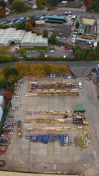 Vertical screen Aerial Timber Yard In Leeds Showing Organized Rows Of Stacked Lumber And Pallets With Coils And Pipes, Forklifts And Small Vehicles Operating On Concrete Yard, Warehouse Buildings