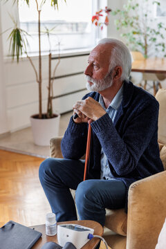 Contemplative senior man is leaning on cane at home