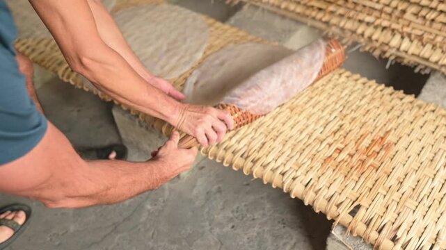 Asian Man Rolling Rice Paper On Mat, Calm Rural Workshop Setting, Hands Skillfully Draw Translucent Sheets From Bamboo Roller, Closeup Of Woven Mat Texture And Stacked Rice Rounds Drying, Warm Natural