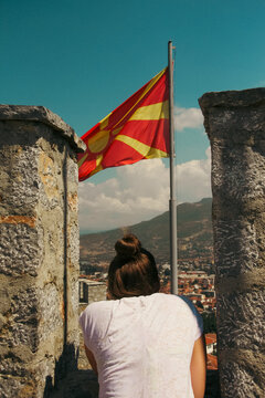 A female traveler stands between the ancient stone walls of Samoil's Fortress overlooking the historic town of Ohrid. The red and yellow national flag of North Macedonia waving in the background