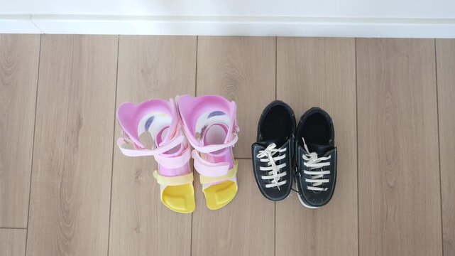 Pair of children's ankle-foot orthosis braces afo in pink and yellow colors standing next to black sneakers on wooden floor in slow motion