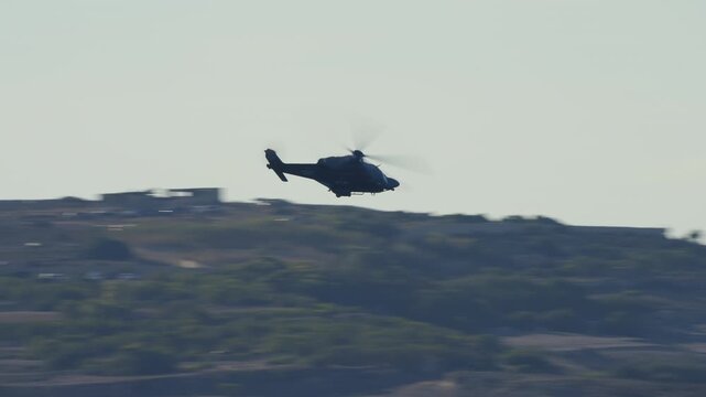 LUQA, MALTA - SEPTEMBER 28, 2025: Video footage of a military-style helicopter silhouetted against a bright, hazy sky. The chopper banks and then flies away over a hilly landscape. Dynamic tracking