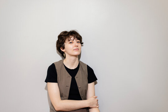Portrait of young woman in vest sitting against plain wall