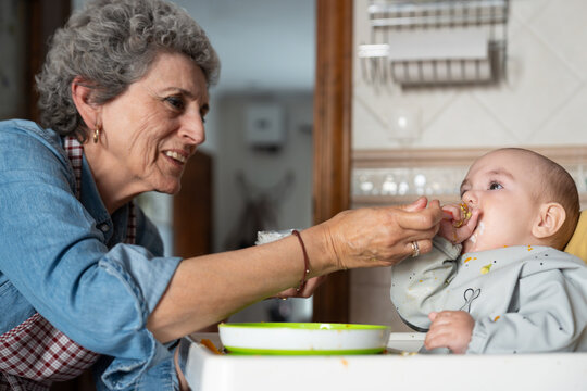 Grandmother feeding baby in high chair with spoon in kitchen