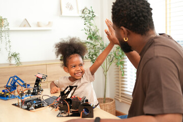 African American father giving high-five to his young daughter after successfully building a DIY robot arm. Concept of STEM education, diversity, and happy family bonding. © Jestercine