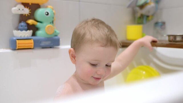 A sweet toddler plays independently during a bath time. A charming little boy smiles happily as he plays with bubble bath and toys during his evening bedtime ritual.